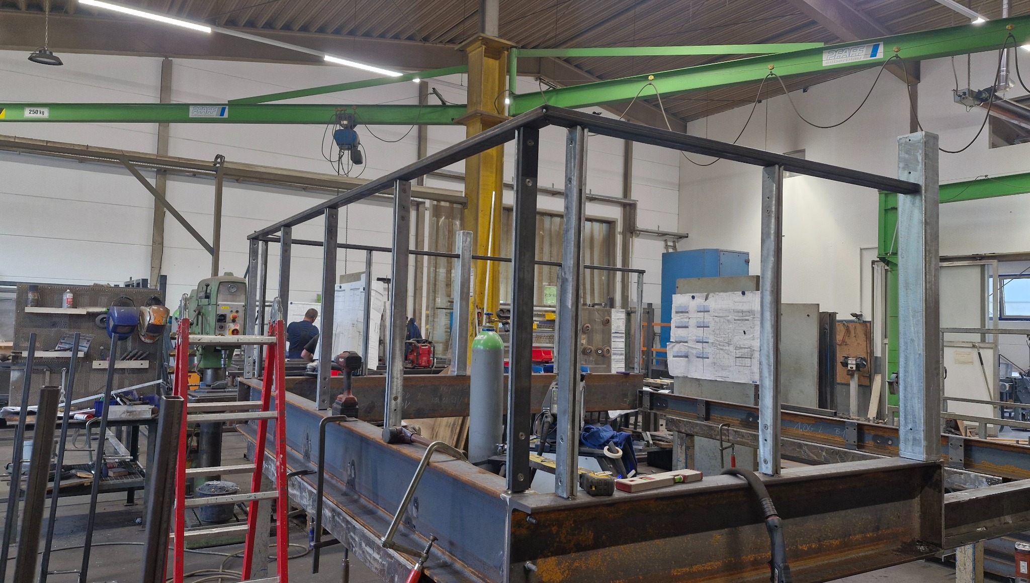 Wnętrze warsztatu z konstrukcją z metalowych profili w trakcie budowy. Widoczny spawacz i narzędzia. Elementy konstrukcyjne w surowym stanie, bez wykończenia.