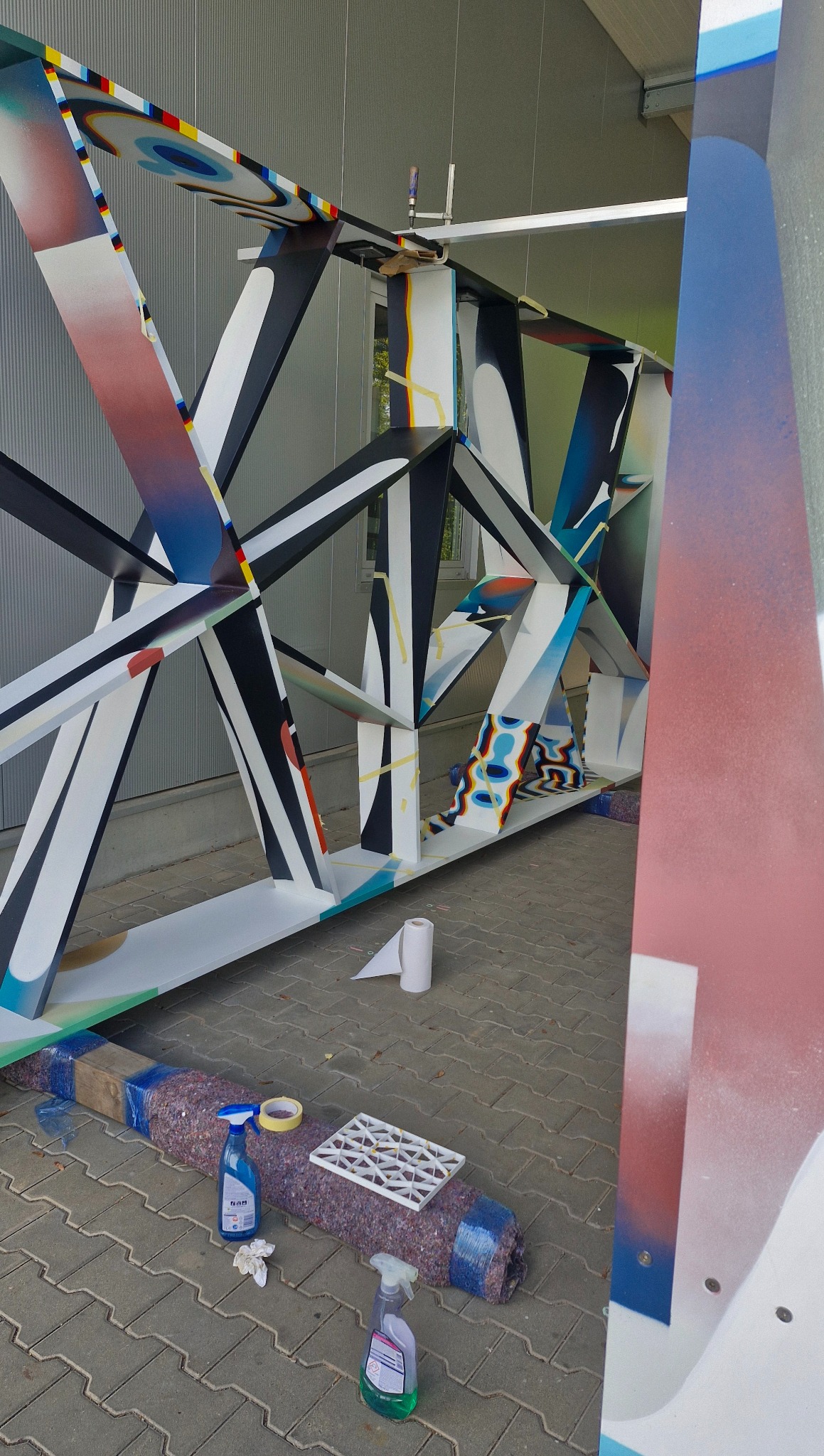 Kolorowa konstrukcja stalowa w trakcie malowania, z widocznymi narzędziami i środkami czystości. Elementy w geometryczne wzory, częściowo zamaskowane.