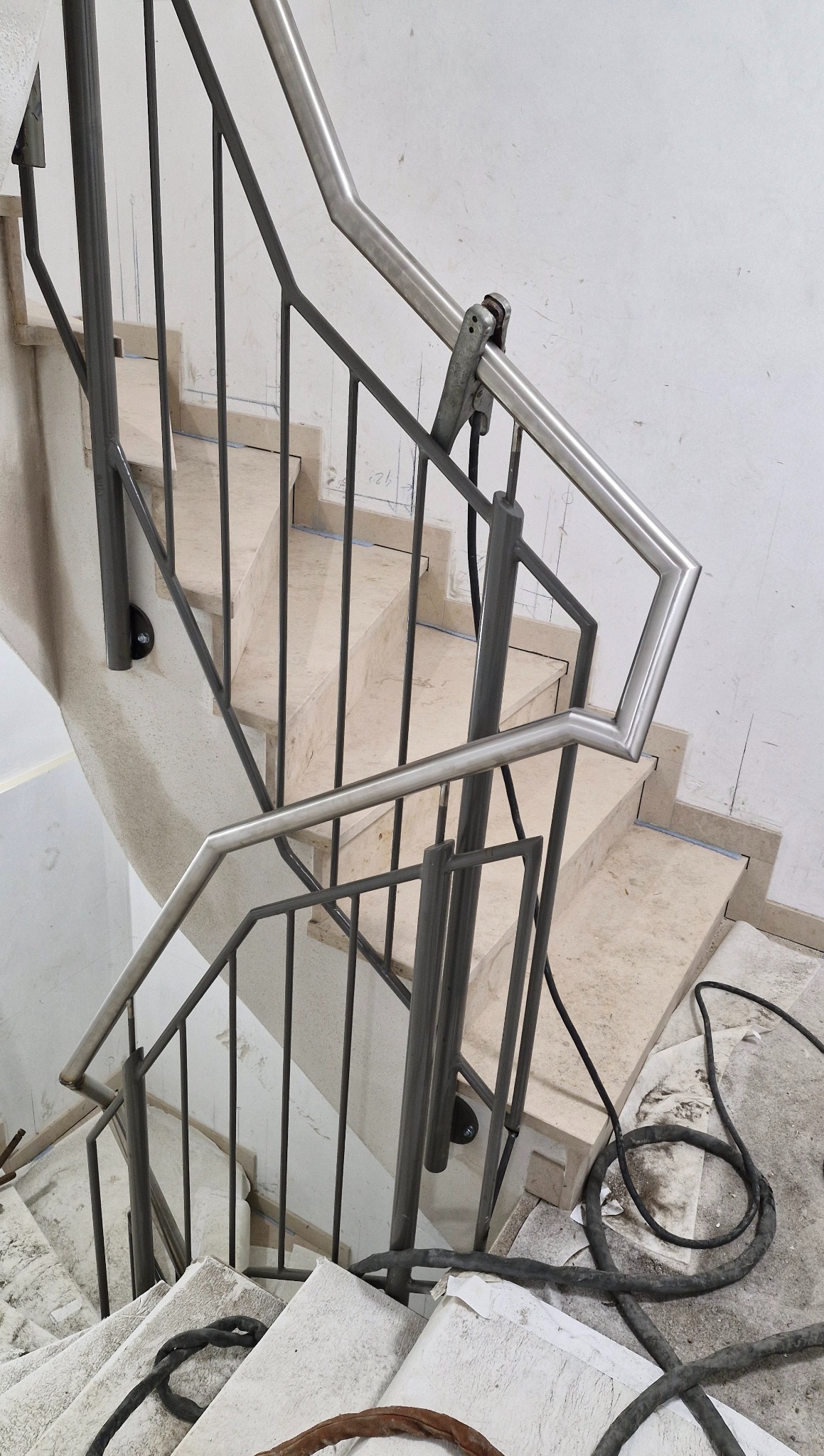 Kątowe ujęcie schodów z metalową balustradą w trakcie montażu. Widoczne przewody i narzędzia, co sugeruje trwający proces instalacji. Jasne, marmurowe stopnie.