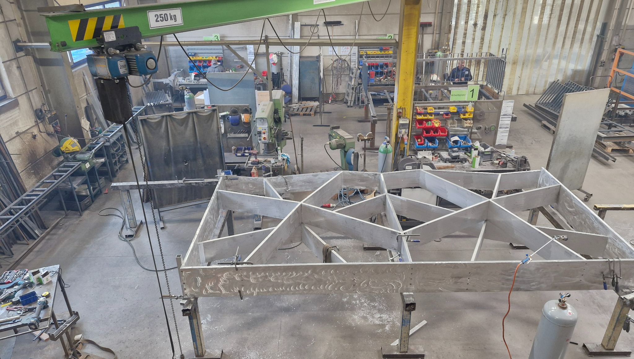 Wnętrze warsztatu z widokiem na metalową konstrukcję szkieletową, prawdopodobnie w trakcie spawania/montażu. W tle widoczne narzędzia i stanowiska pracy.