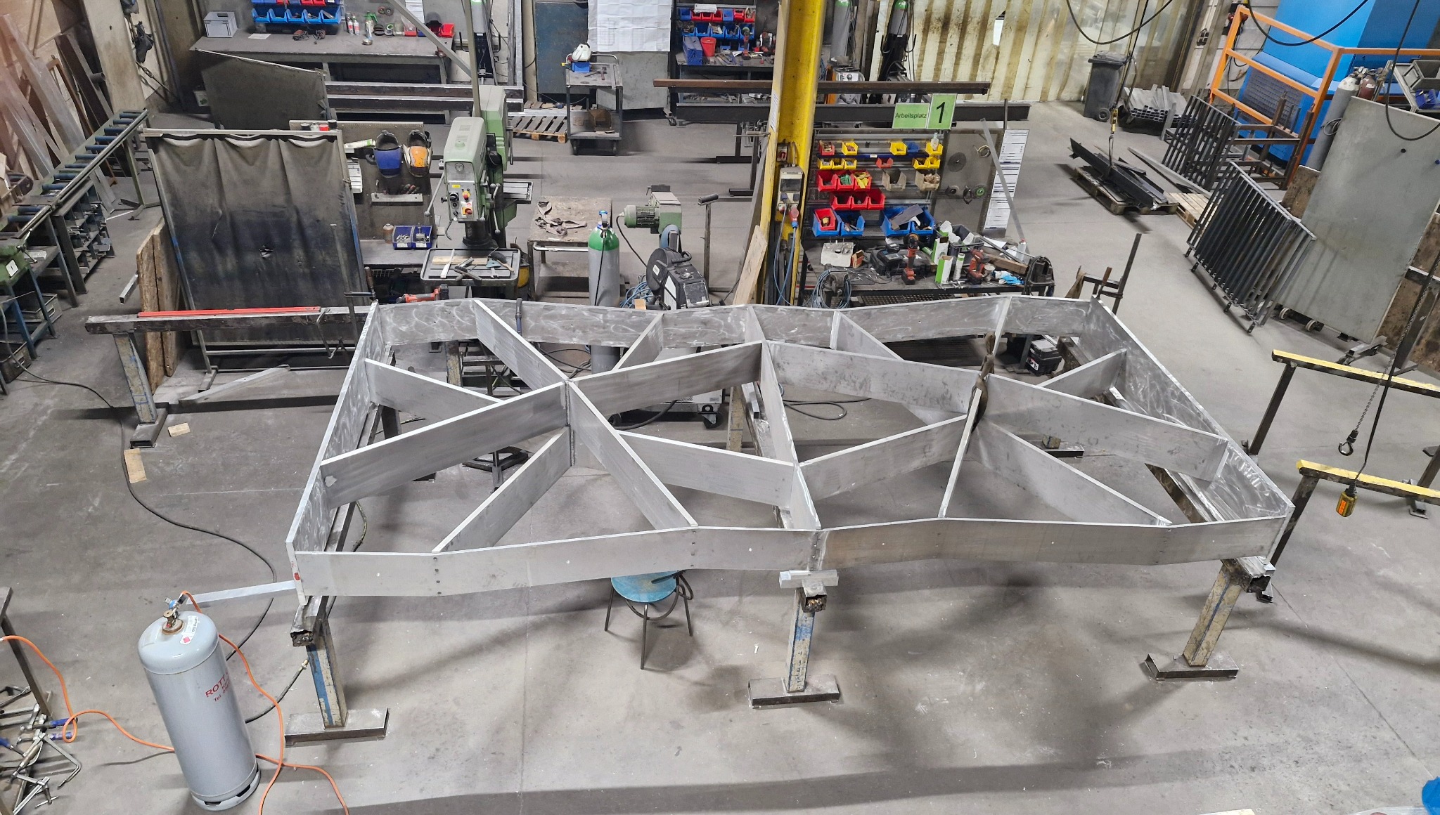 Metalowa konstrukcja szkieletowa z profili, spawana, na stojakach w hali produkcyjnej. Widok z góry, butla z gazem w lewym dolnym rogu.