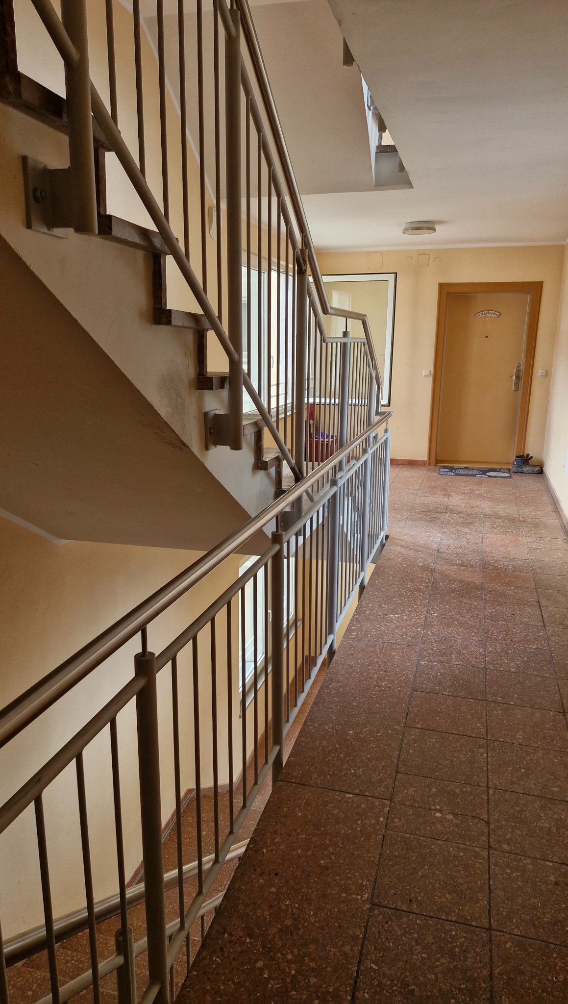 Klatka schodowa w bloku z metalowymi, pionowymi barierkami. Widok z dołu na schody i poręcze. Jasne ściany i brązowa podłoga z płytek.