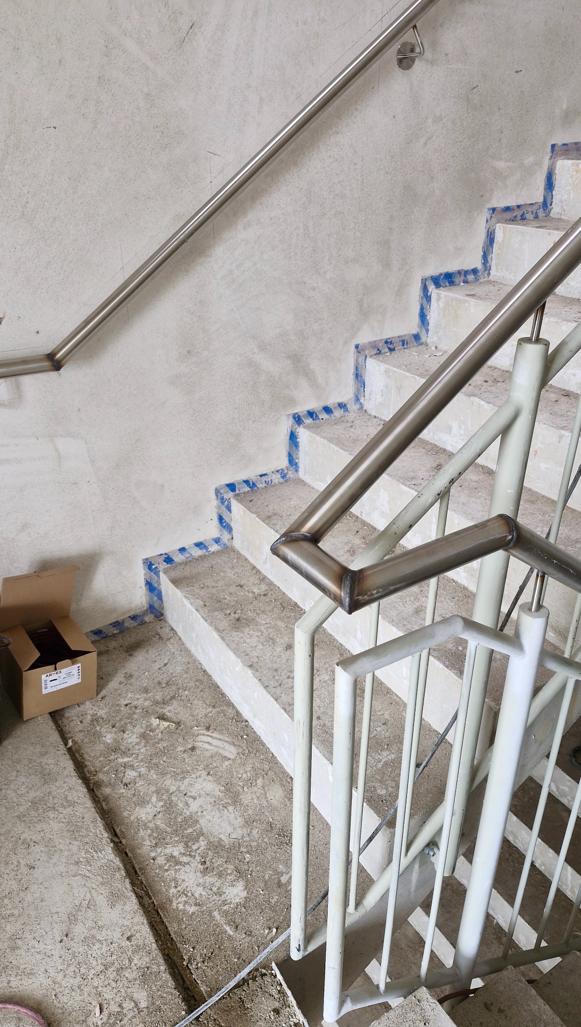 Metalowe poręcze i balustrady na betonowych schodach w trakcie montażu, widoczne zabezpieczenia na stopniach. Karton na podłodze. Ujęcie z góry.