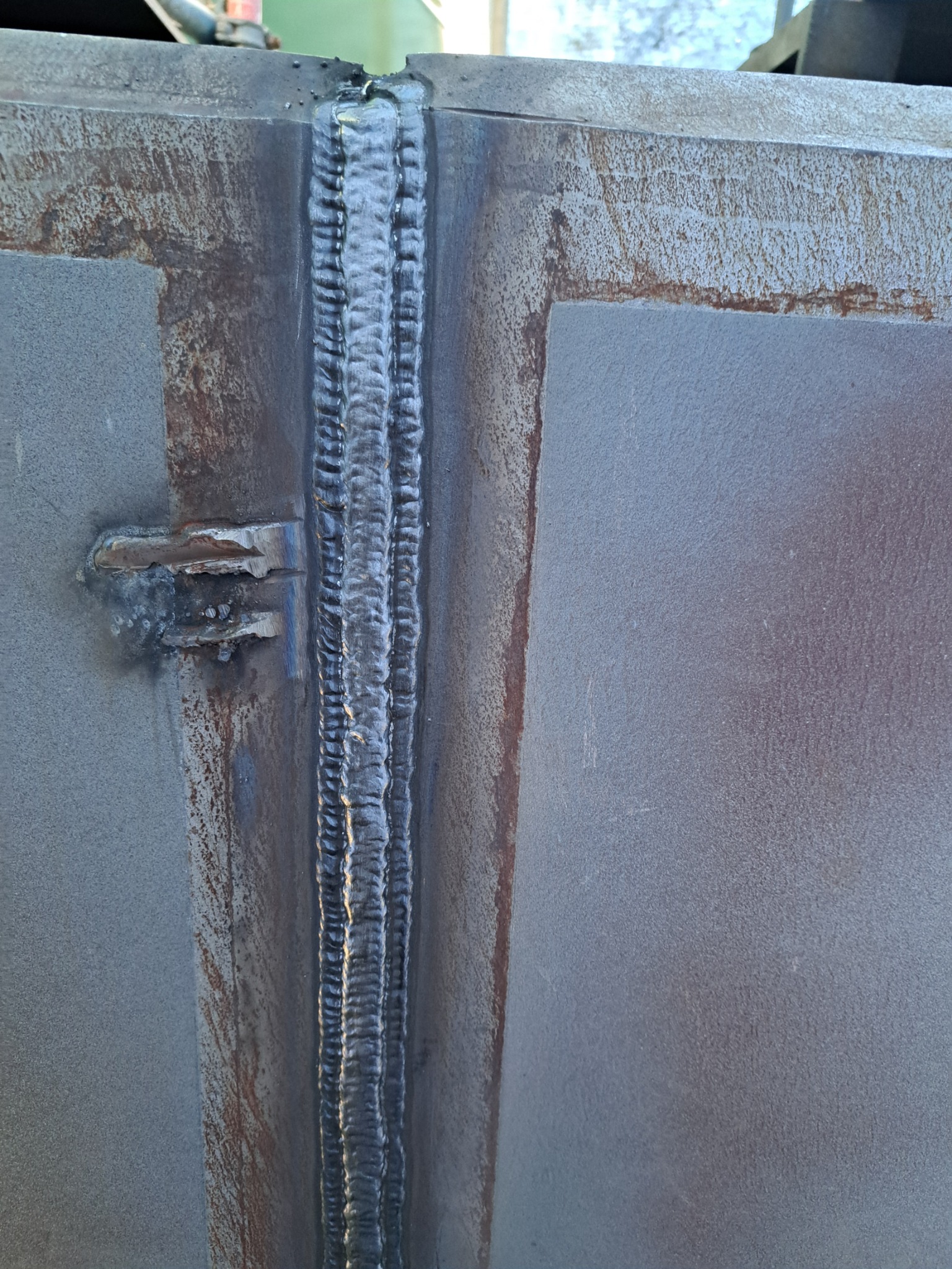 Szlifowany spaw na styku dwóch metalowych płyt, widoczne ślady rdzy i niedoskonałości obróbki, surowy industrialny charakter, zbliżenie na spoinę.