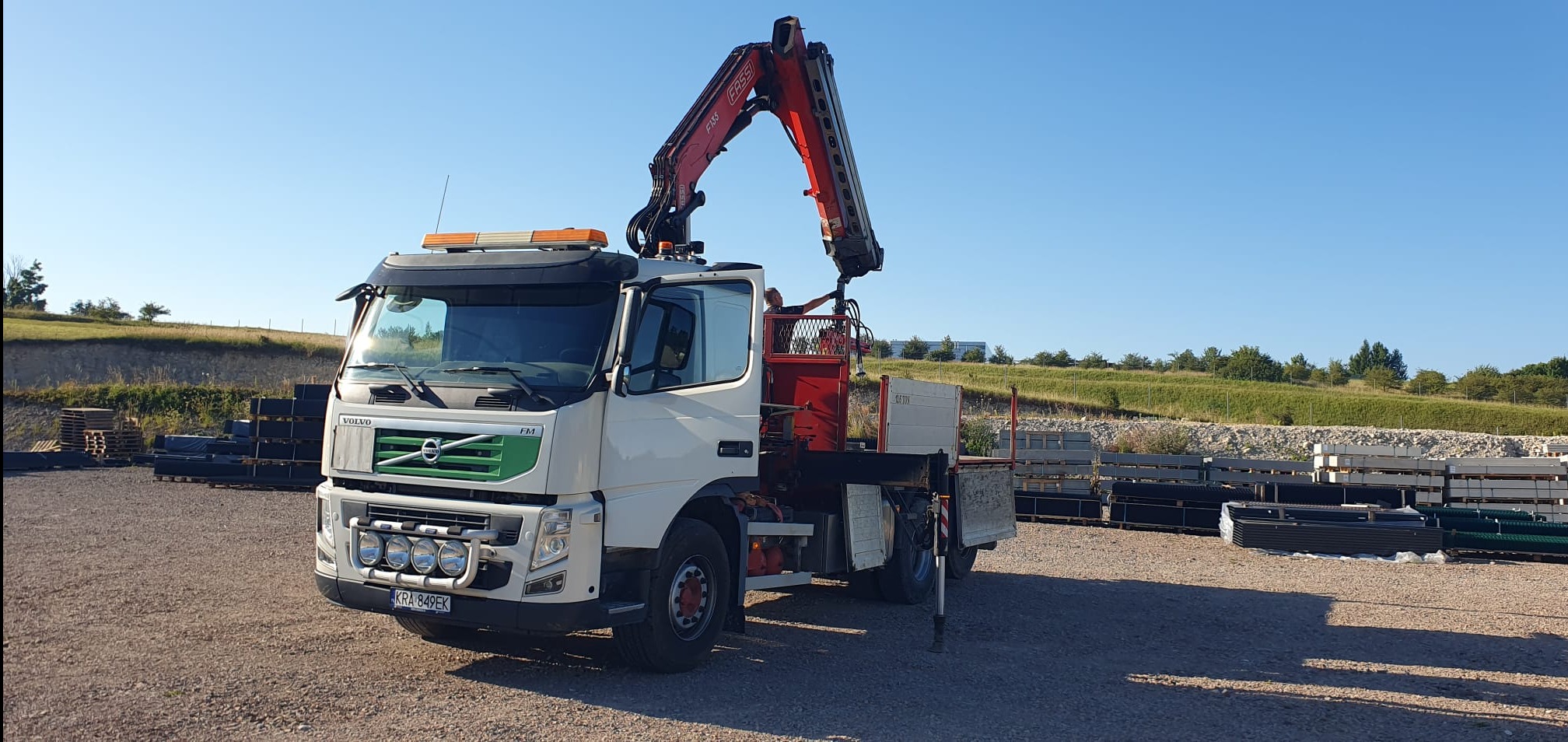 Biała ciężarówka Volvo FM z dźwigiem Fassi F135 na żwirowym placu, z ułożonymi elementami konstrukcyjnymi w tle, w słoneczny dzień.