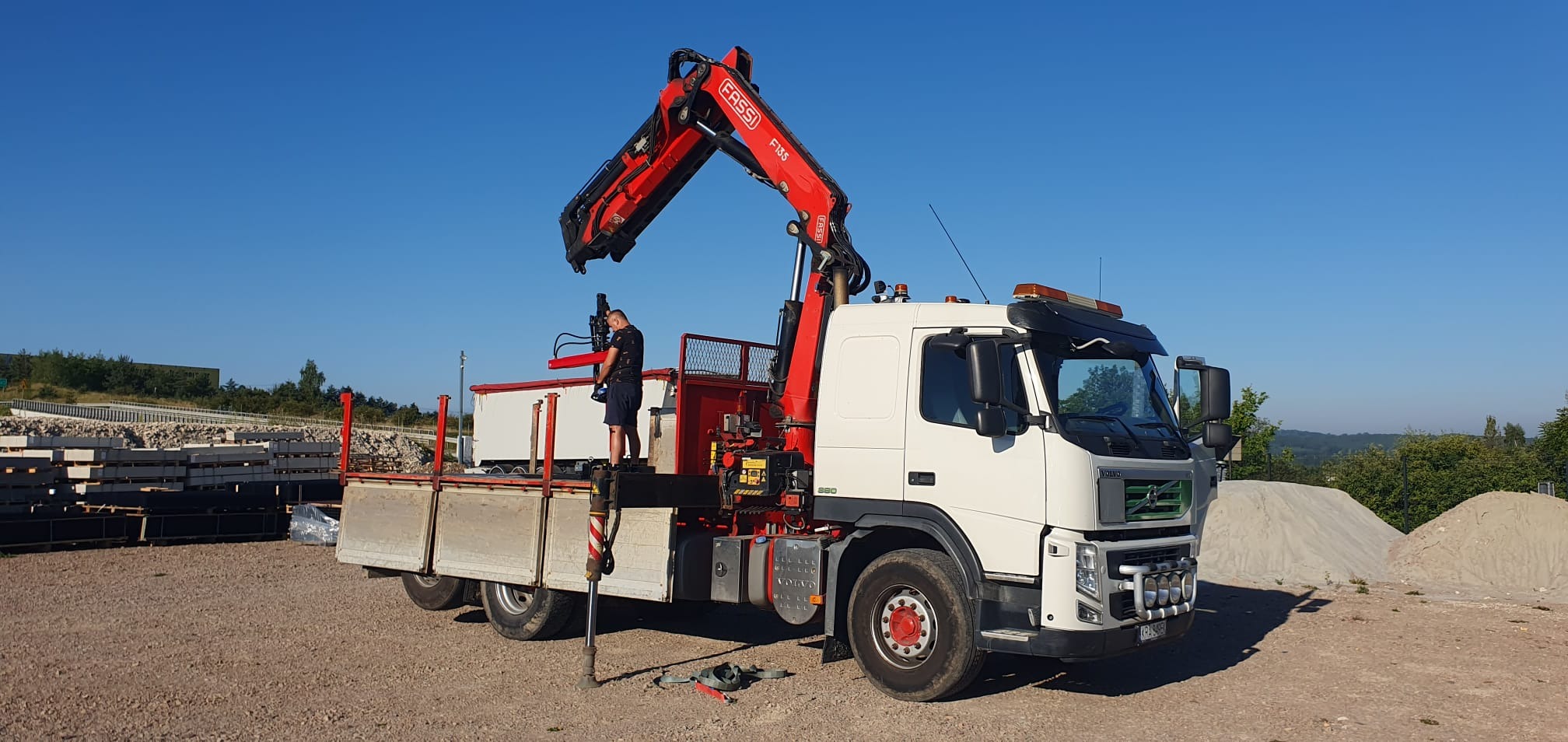 Ciężarówka Volvo z dźwigiem Fassi F135 załadowana betonowymi elementami. Operator w trakcie pracy na placu budowy. W tle hałda piachu i zieleń.