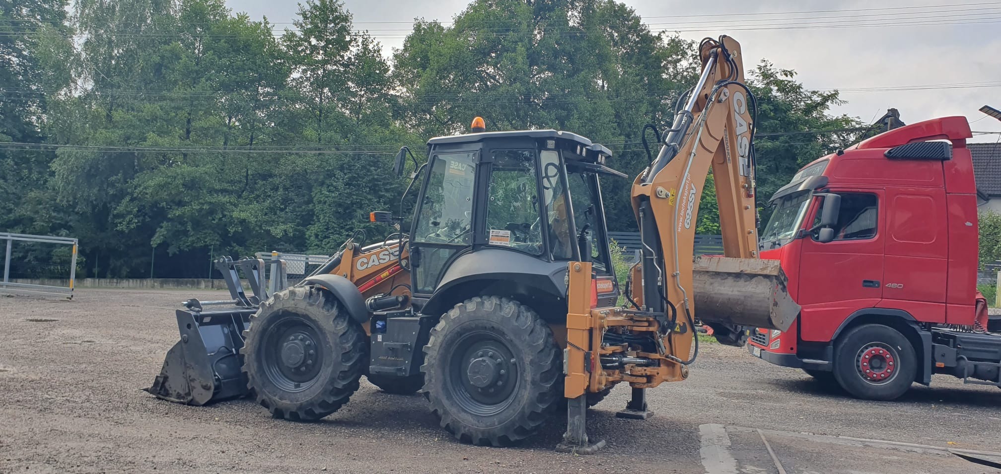 Pomarańczowo-czarna koparko-ładowarka CASE na żwirowym terenie, z podniesioną łyżką i widoczną czerwoną ciężarówką w tle. Maszyna gotowa do pracy.