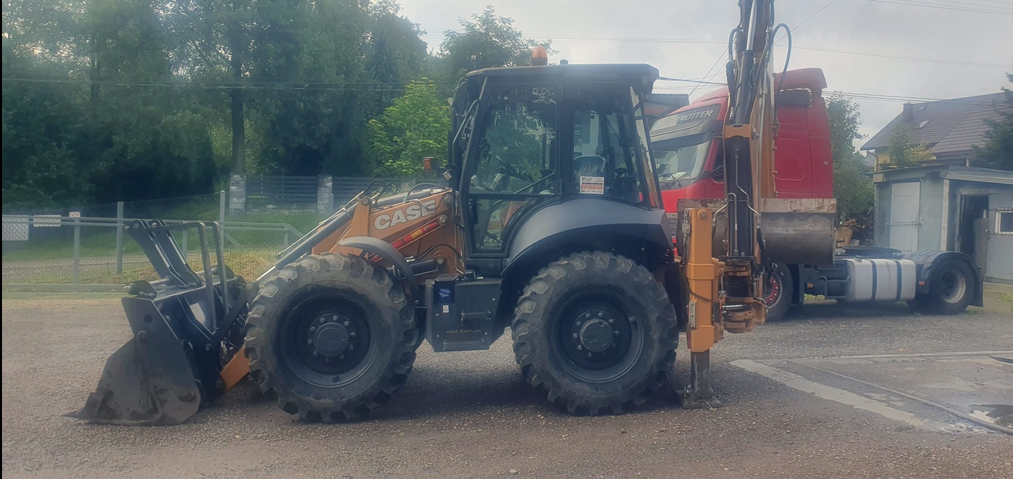 Pomarańczowo-czarna koparko-ładowarka Case z łyżką i wysięgnikiem na tle czerwonej ciężarówki i zieleni. Maszyna stoi na żwirowym podłożu, gotowa do pracy.