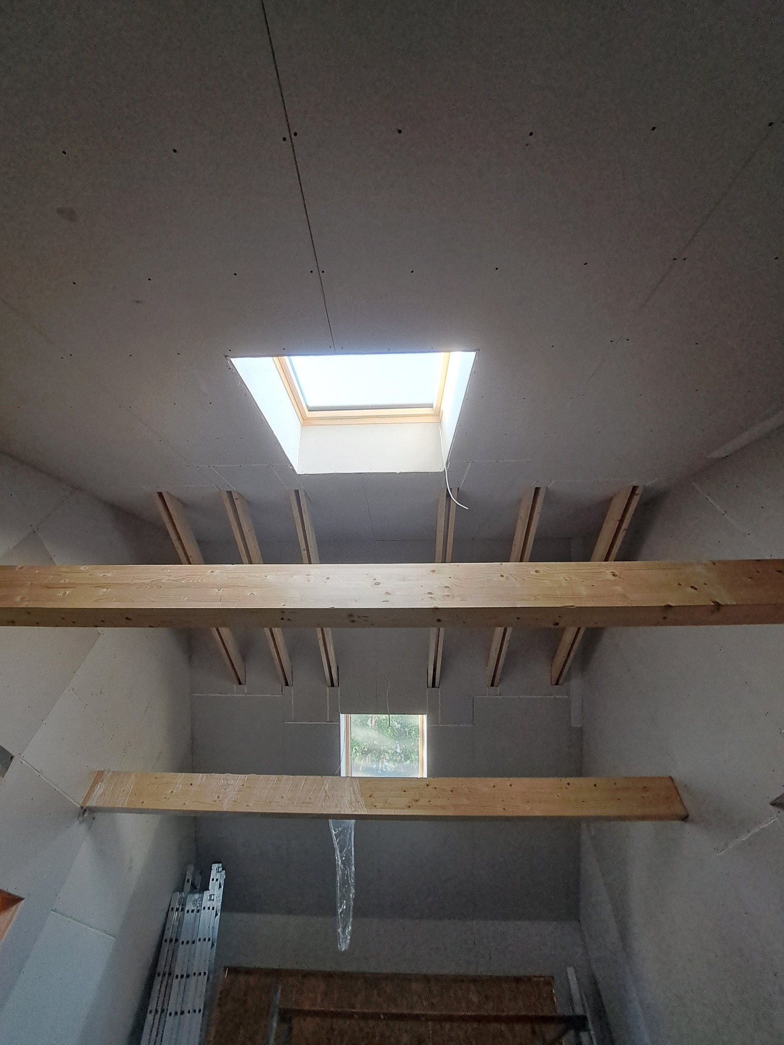 Montaż płyt karton-gips na poddaszu z oknem dachowym i drewnianą konstrukcją wsporczą. Widoczna drabina i folia zabezpieczająca.