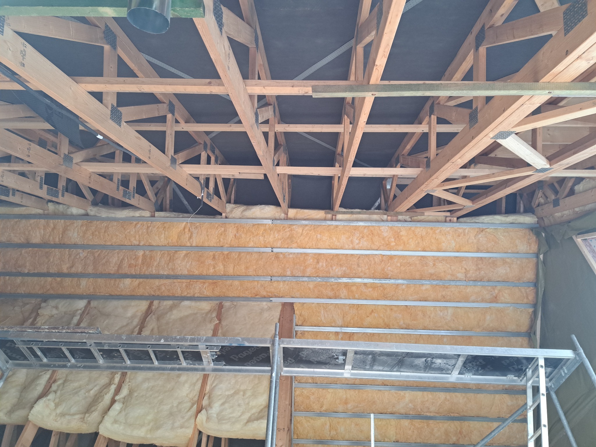 Ocieplenie poddasza wełną mineralną, widoczne drewniane elementy konstrukcyjne dachu i metalowy ruszt. Prace na wysokości, drabina.