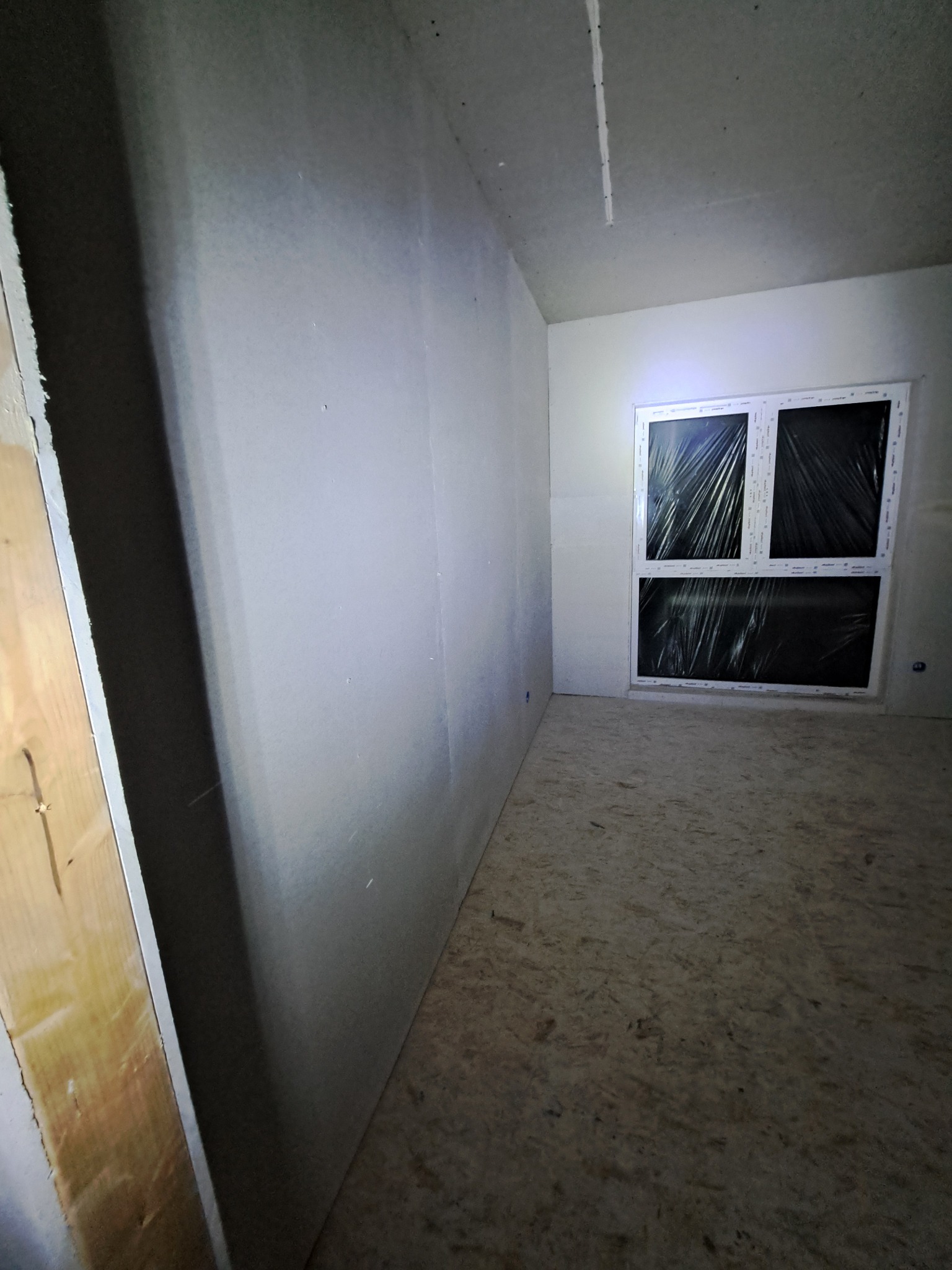 Pokój w trakcie prac wykończeniowych: ściany z płyt kartonowo-gipsowych, zabezpieczone okno folią, surowa podłoga z płyt OSB. Widok na narożnik pomieszczenia.