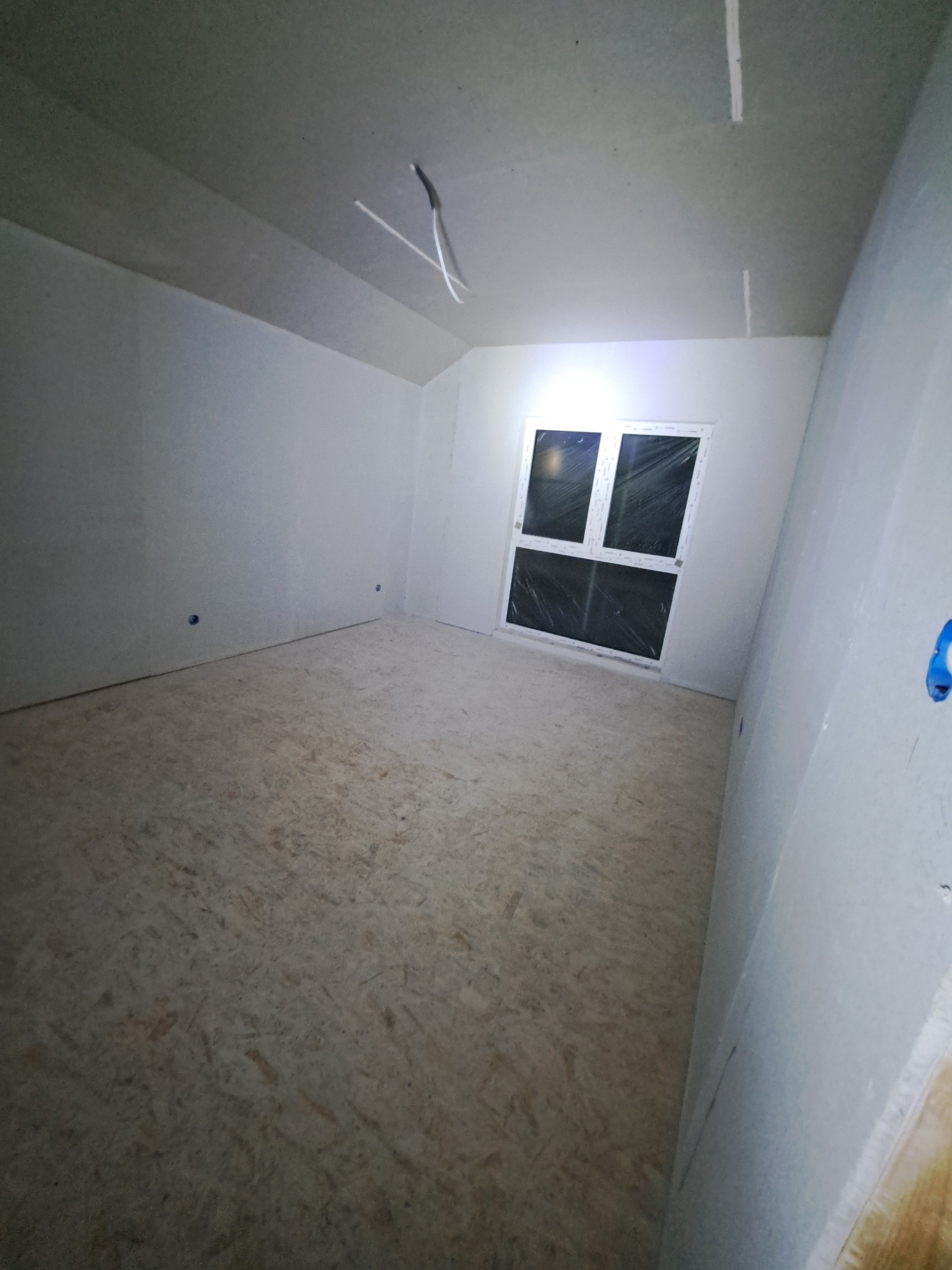 Pokój na poddaszu z oknem, ściany wykończone płytami gipsowo-kartonowymi, widoczne łączenia płyt. Podłoga wyłożona płytami OSB. Surowy stan wykończenia.