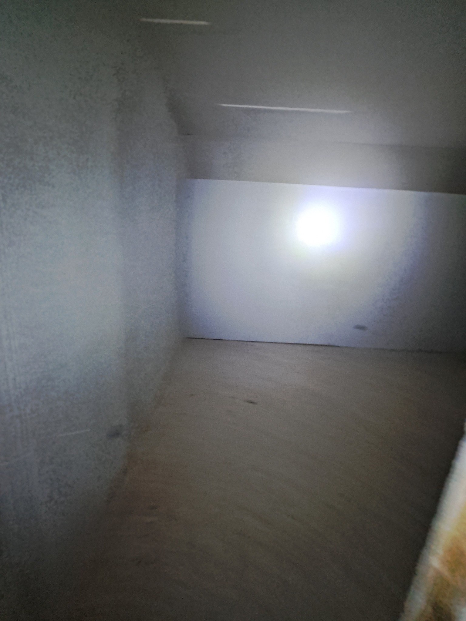 Surowe wnętrze pokoju w trakcie prac wykończeniowych. Ściany i sufit pokryte świeżym tynkiem, podłoga przygotowana do dalszych prac. Oświetlenie punktowe.