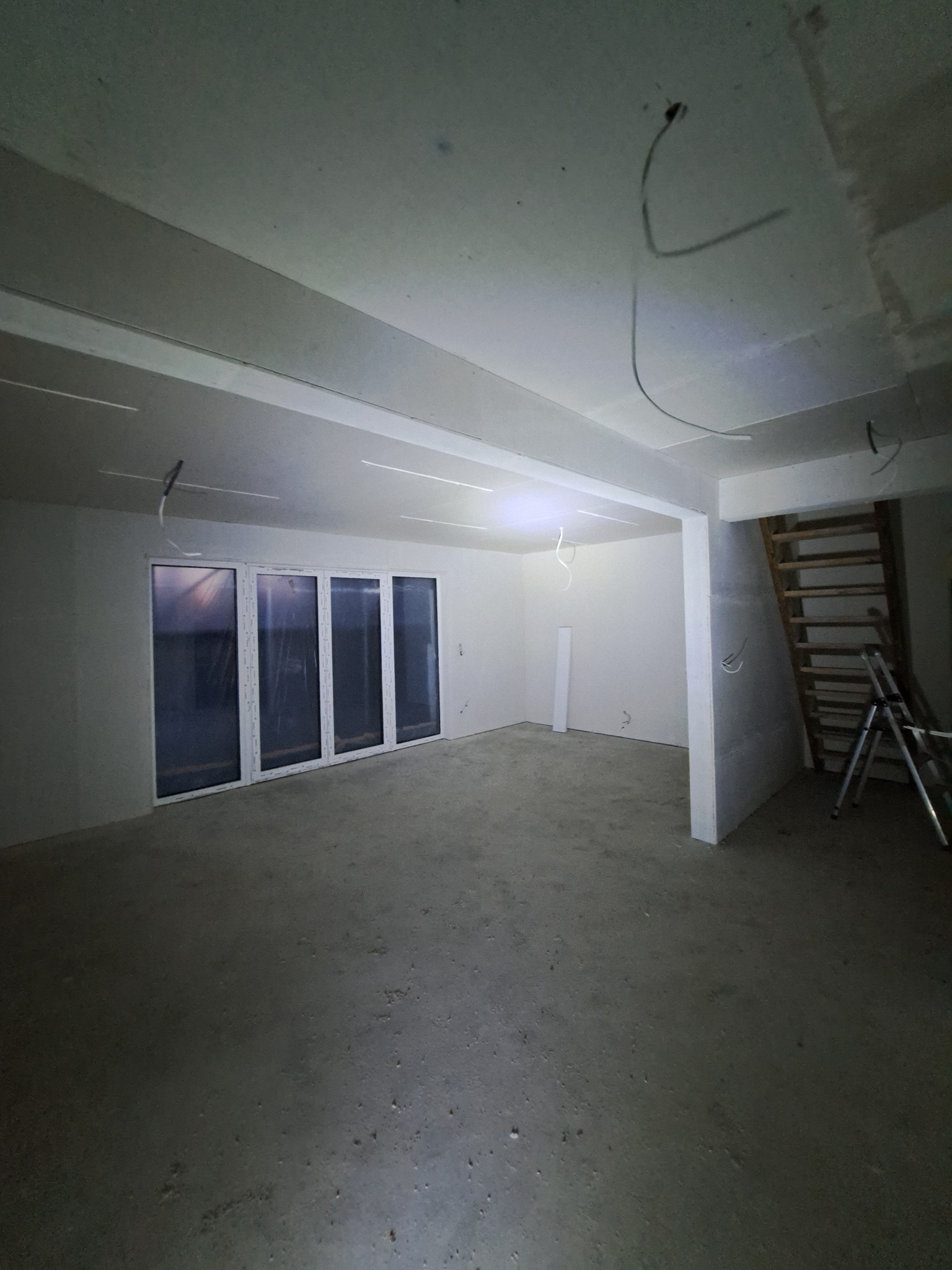 Surowe wnętrze w trakcie prac wykończeniowych: białe ściany, okna PCV, betonowa podłoga, widoczne przewody elektryczne i drabina. Etap przygotowania do dalszych prac.