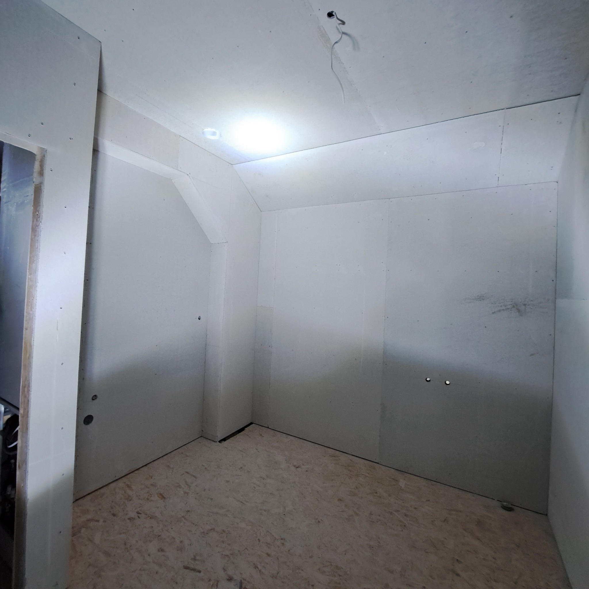 Wnętrze w trakcie prac wykończeniowych: ściany obłożone płytami gipsowo-kartonowymi, widoczne łączenia i otwory instalacyjne, podłoga z płyt OSB. Oświetlenie punktowe.