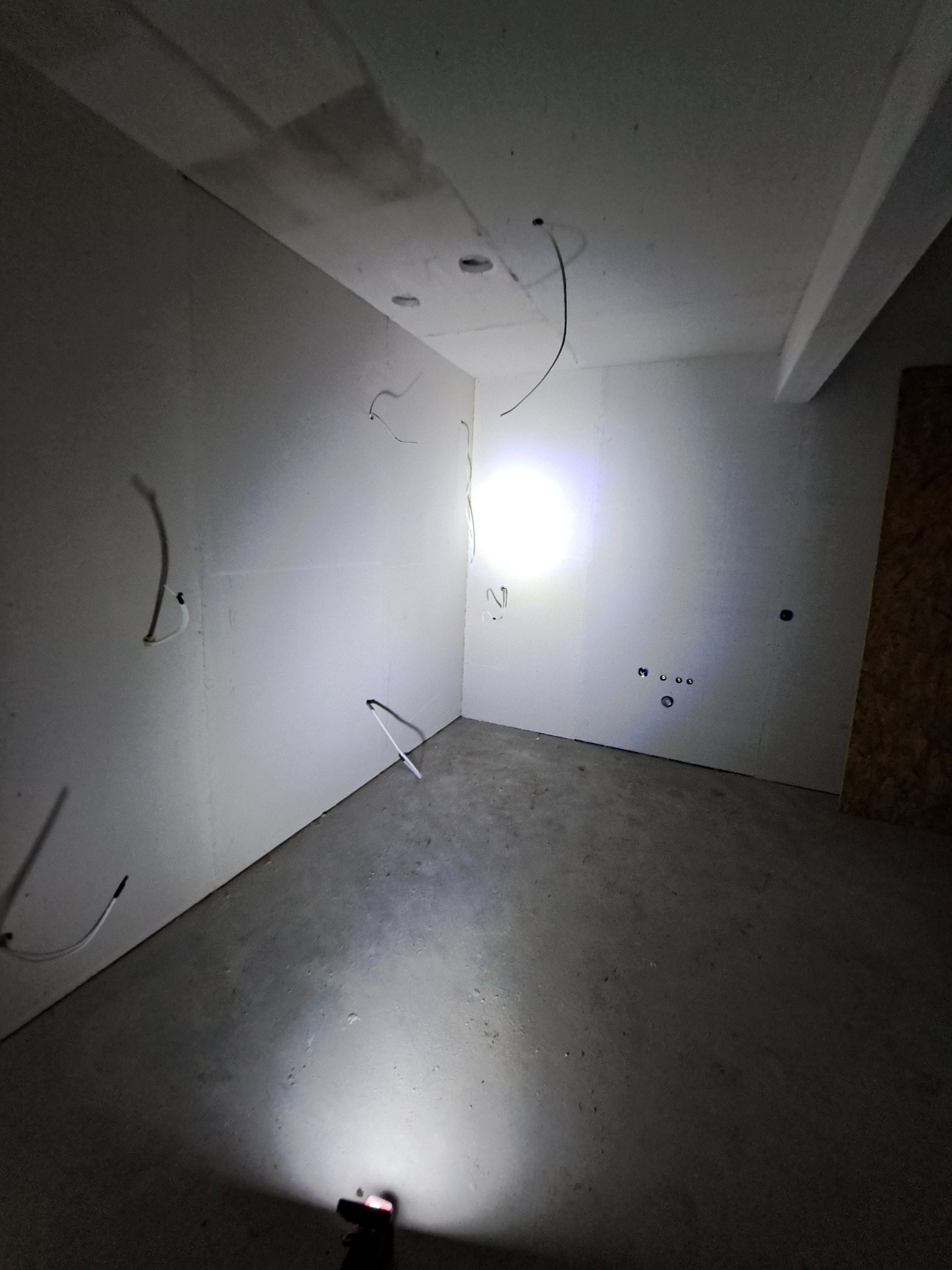 Surowe wnętrze z płytami gipsowo-kartonowymi na ścianach i wystającymi przewodami elektrycznymi. Widoczne otwory na oświetlenie w suficie i gniazdka na ścianie.