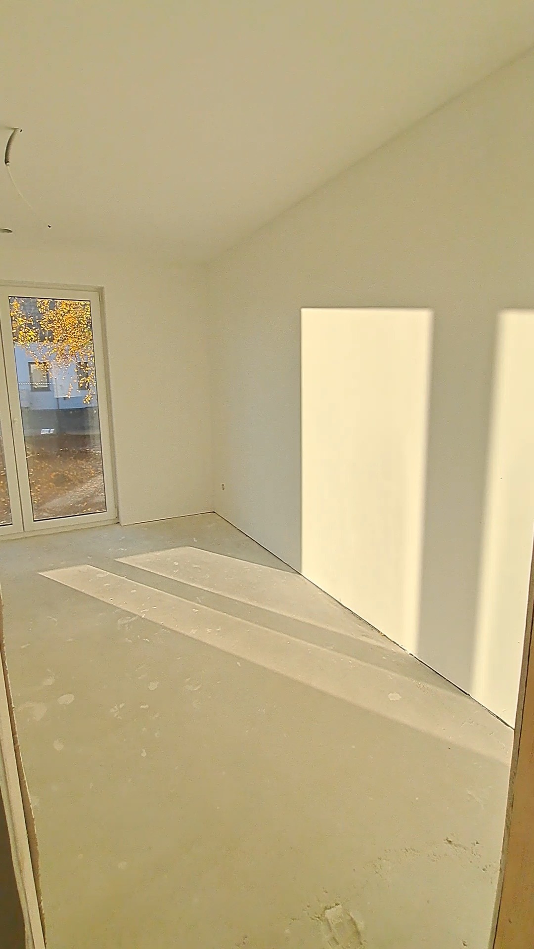 Pusty pokój po malowaniu ścian na biało, z widocznym oknem i betonową podłogą. Świeże, jasne wnętrze w trakcie wykańczania, z promieniami słońca wpadającymi przez okno.