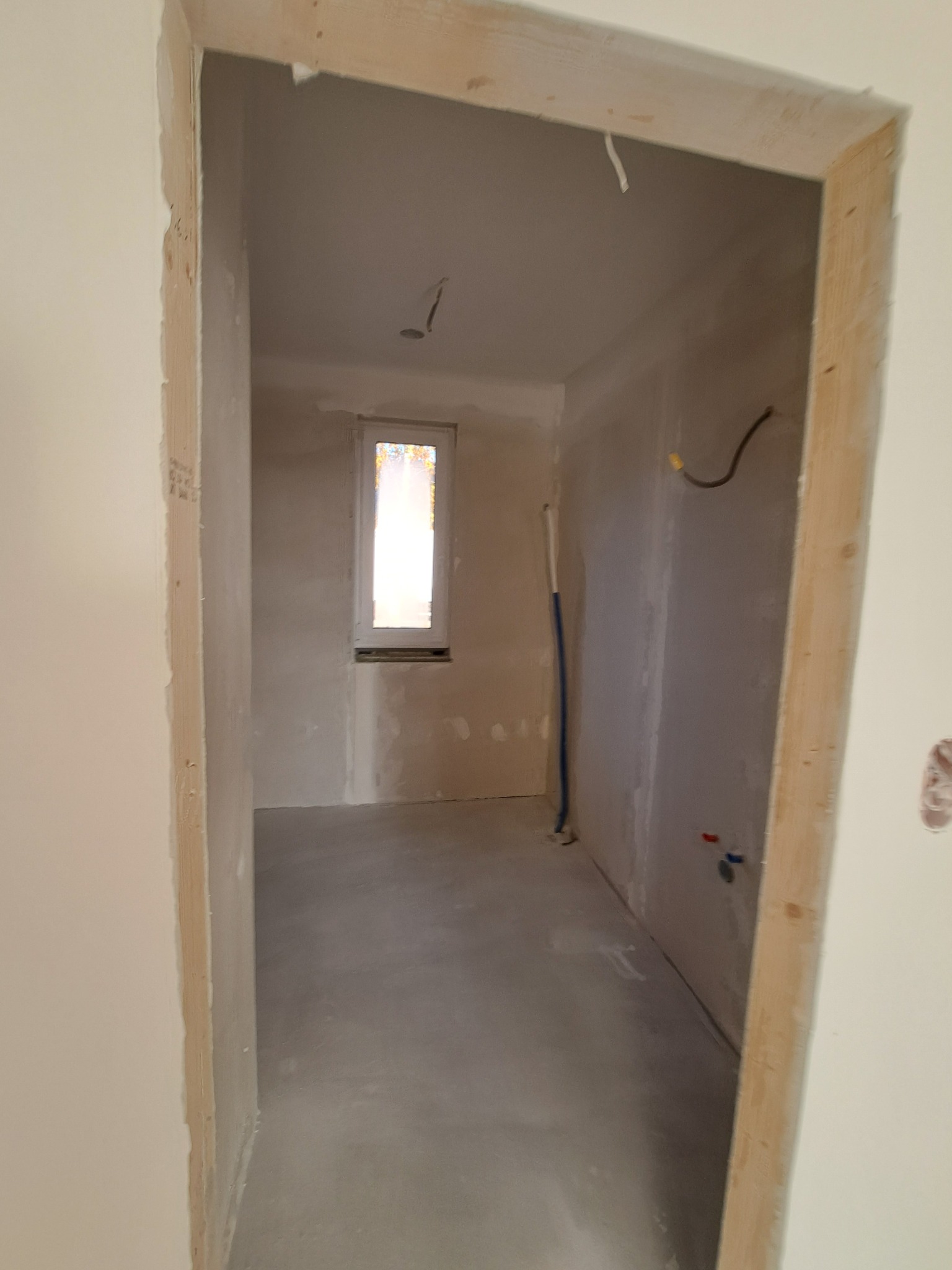 Surowe wnętrze w trakcie prac wykończeniowych. Widoczne ściany po gładzi, surowa ościeżnica i instalacje. Okno z widokiem na zewnątrz. Podłoga wylewka.