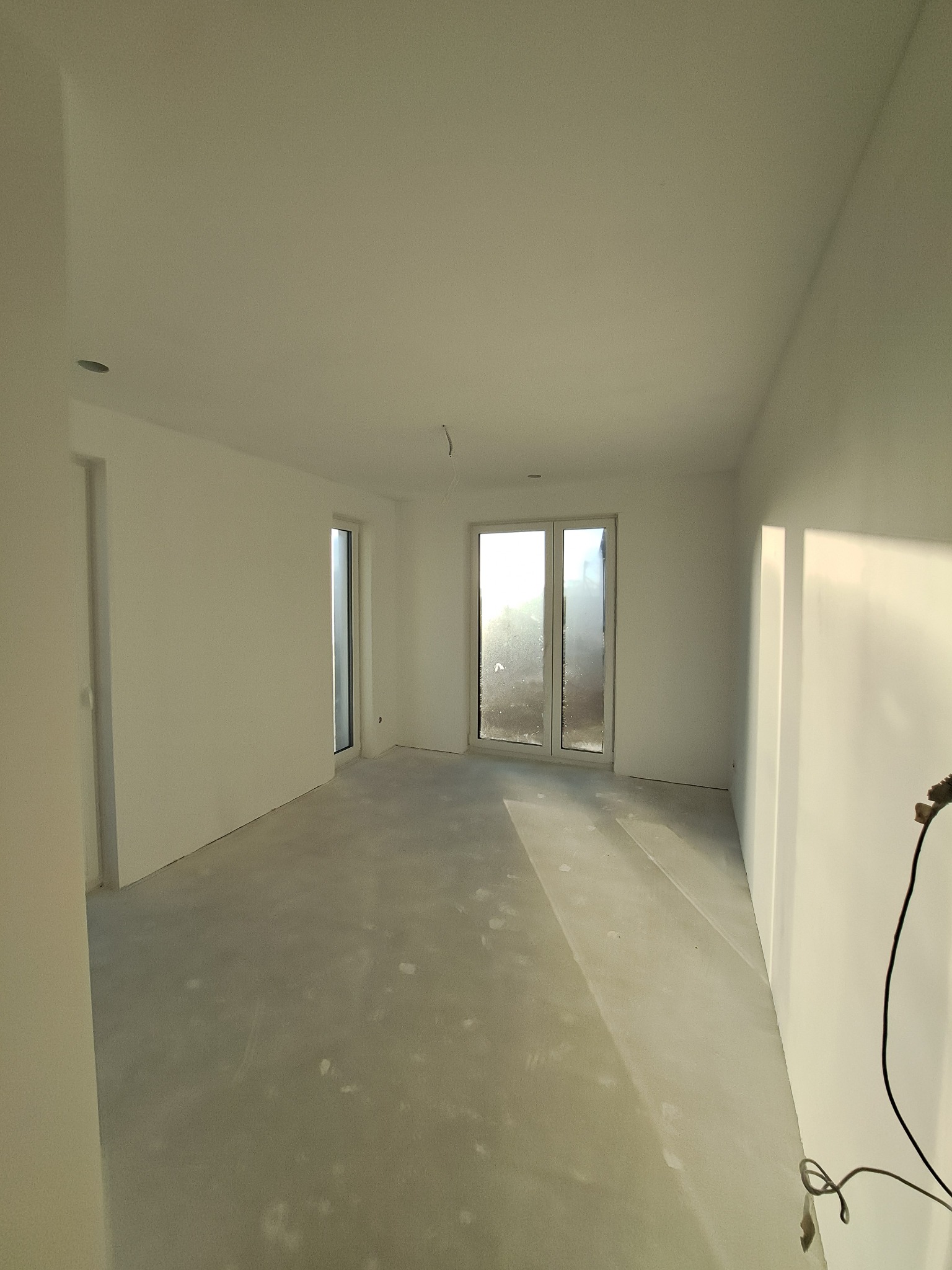 Jasne, puste wnętrze po remoncie z białymi ścianami, betonową podłogą i dużymi oknami balkonowymi wpuszczającymi światło. Widoczne ślady po pracach wykończeniowych.