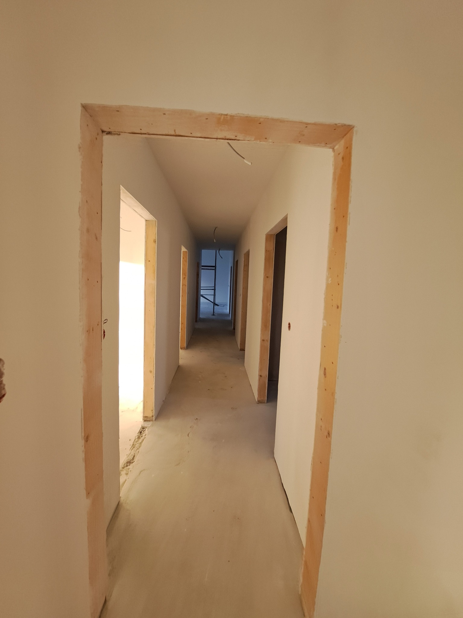 Korytarz w trakcie prac wykończeniowych. Widoczne surowe ościeżnice drzwiowe i świeżo pomalowane ściany. W tle rusztowanie.