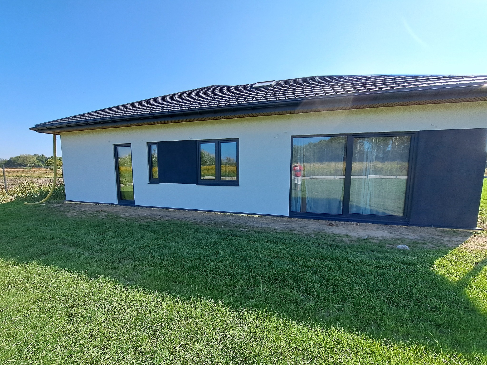 Nowoczesny dom jednorodzinny z białymi ścianami i antracytowymi akcentami, widok z zewnątrz na tle zielonego trawnika i błękitnego nieba. Widoczne okna i drzwi.