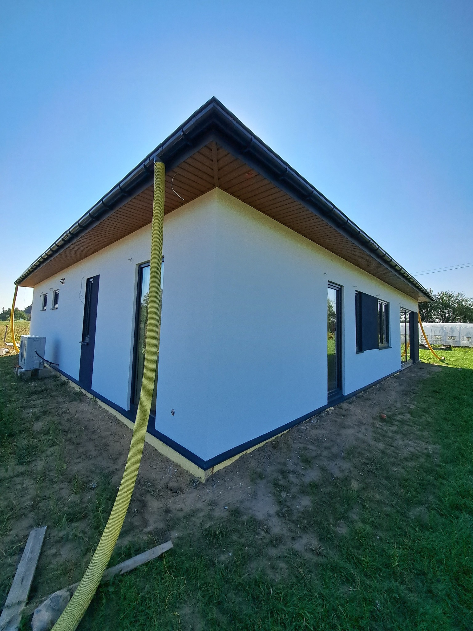 Nowoczesny, parterowy dom po remoncie elewacji, z widoczną żółtą rurą wentylacyjną i klimatyzatorem. Świeżo pomalowana fasada w jasnym odcieniu, ciemne okna i dach.