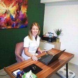Urszula Banaszek dietetyk kliniczny, psychodietetyk - Dietetyk w gabinecie: Kobieta w białym uniformie pracuje przy biurku z laptopem, w tle waga, obraz z motylem i roślina doniczkowa.