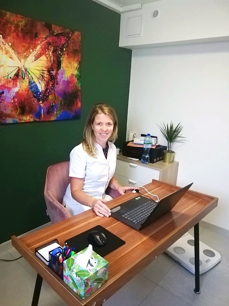 Dietetyk w gabinecie: Kobieta w białym uniformie pracuje przy biurku z laptopem, w tle waga, obraz z motylem i roślina doniczkowa.