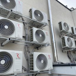 JERZY FRANK HYDROPROJEKT - Ściana budynku z zamontowanymi klimatyzatorami Rotenso, widoczne przewody i metalowe konstrukcje wsporcze. Ujęcie z dołu, podkreślające skalę instalacji.