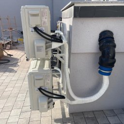JERZY FRANK HYDROPROJEKT - Zewnętrzne jednostki klimatyzacji zamontowane na elewacji budynku, z widocznymi przewodami i elementami instalacji. Widoczne narzędzia i materiały budowlane w tle.
