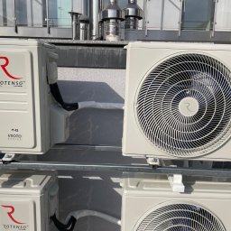 JERZY FRANK HYDROPROJEKT - Zewnętrzne jednostki klimatyzacji Rotenso zamontowane na metalowej konstrukcji przy ścianie budynku. Widoczne rury i przewody. Na dachu kominy wentylacyjne.