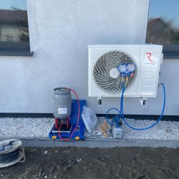 JERZY FRANK HYDROPROJEKT - Biała jednostka zewnętrzna klimatyzacji Rotenso, podłączona do manometrów i butli z czynnikiem chłodniczym, podczas serwisu na tle szarej elewacji domu.