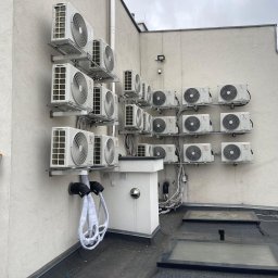 JERZY FRANK HYDROPROJEKT - Rząd klimatyzatorów na elewacji budynku, zamontowanych na metalowych wspornikach. Widoczne kable i rury. Na pierwszym planie drabina i fragment osoby.