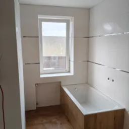 Biała łazienka z drewnopodobną obudową wanny i podłogą. Ściany wyłożone kafelkami z dekoracyjnymi listwami. Widoczne okno z widokiem na zewnątrz.