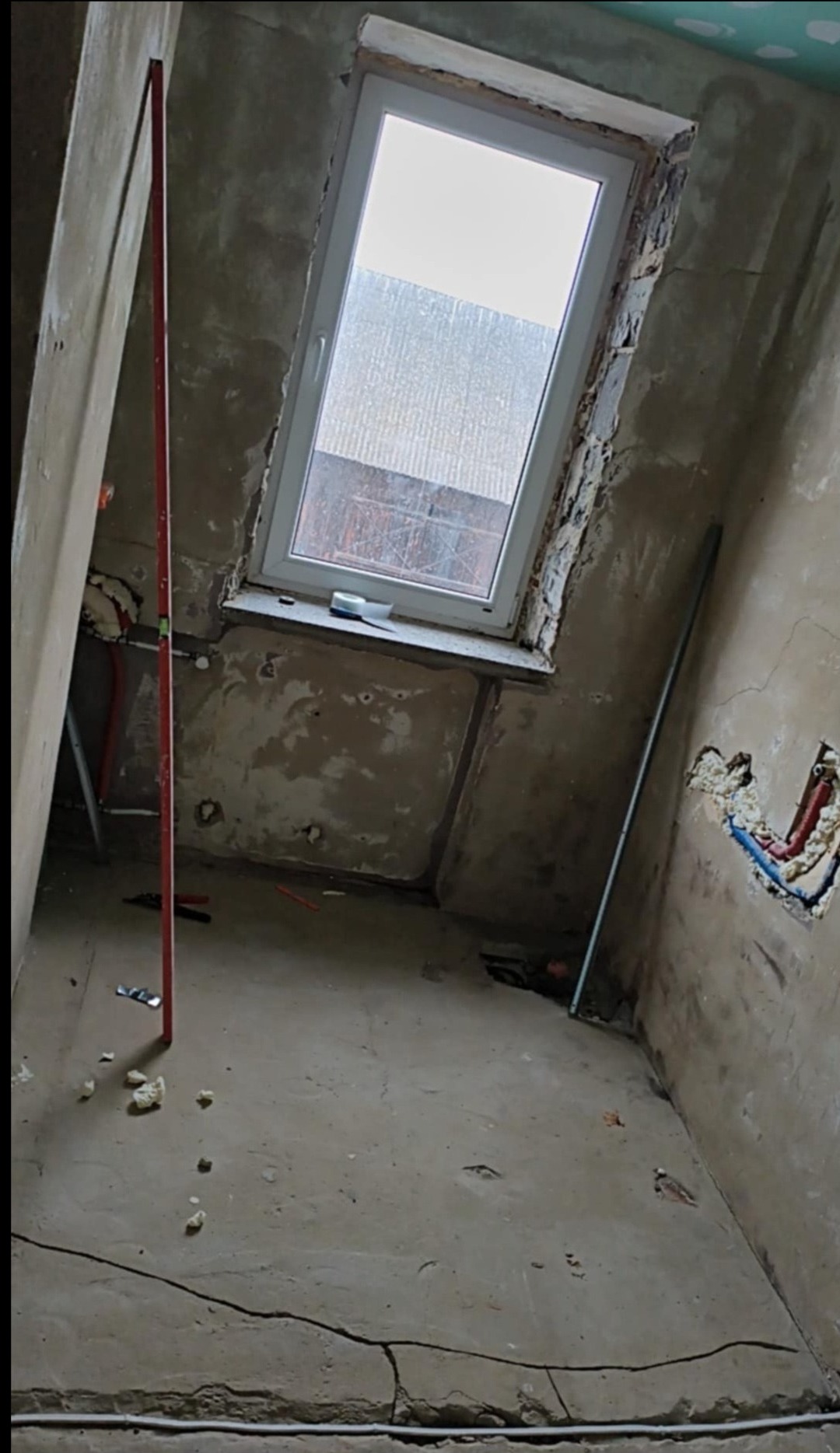 Wnętrze w trakcie remontu: surowe ściany, okno dachowe, widoczne instalacje. Poziomica pionowa przy ścianie, pianka montażowa na podłodze.