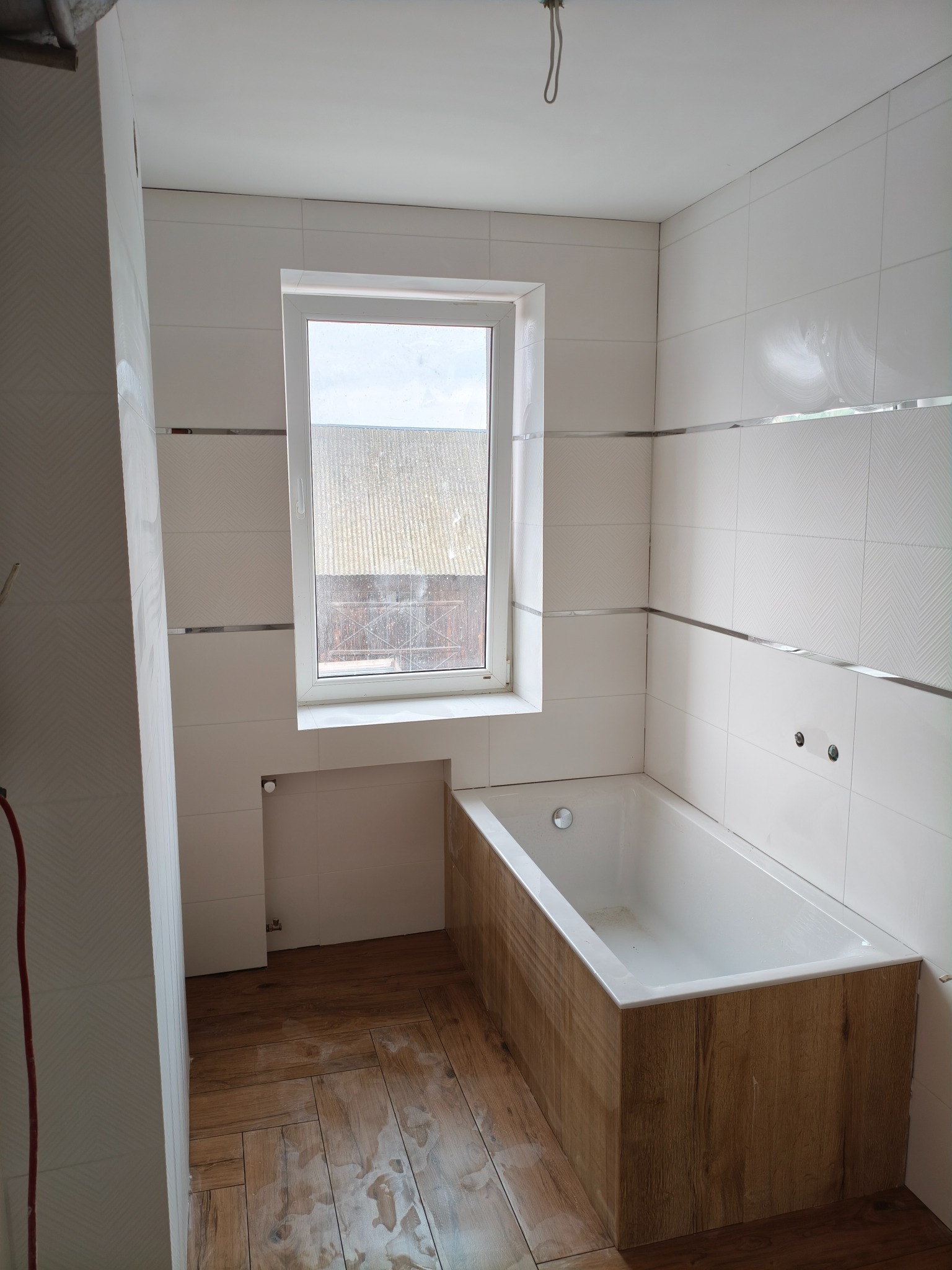 Biała łazienka z drewnopodobną obudową wanny i podłogą. Ściany wyłożone kafelkami z dekoracyjnymi listwami. Widoczne okno z widokiem na zewnątrz.