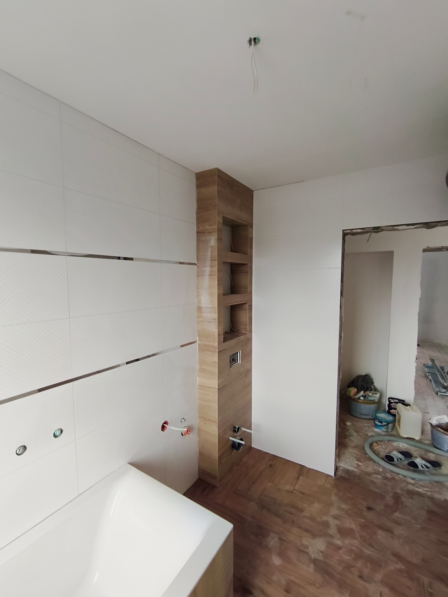 Wnętrze łazienki w trakcie prac wykończeniowych: białe płytki z dekoracyjnym pasem, drewnopodobna zabudowa z półkami i fragment wanny. Podłoga wyłożona panelami.