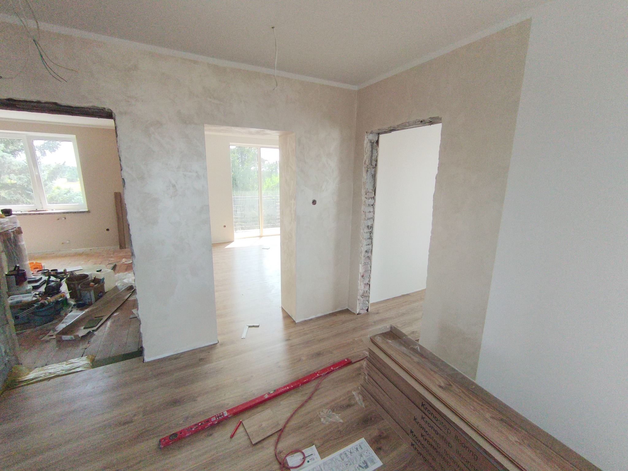 Wnętrze w trakcie prac wykończeniowych: ułożone panele podłogowe, ściany po gładziowaniu z widocznymi otworami drzwiowymi, narzędzia pracy i materiały budowlane.