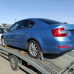 Błękitna Skoda Octavia zabezpieczona pasami na platformie lawety, widok z tyłu, słoneczny dzień.