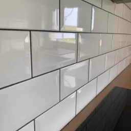 Błyszczące, białe płytki typu metro z czarną fugą ułożone na ścianie w kuchni. Widoczny fragment blatu i płyty indukcyjnej. Refleksy światła na powierzchni płytek.