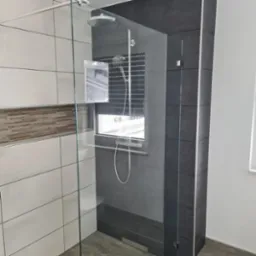 Nowoczesna kabina prysznicowa z deszczownicą, wykończona białymi i grafitowymi płytkami. Na ścianie dekoracyjny pas mozaiki. Widok z wnętrza łazienki, okno z roletą.