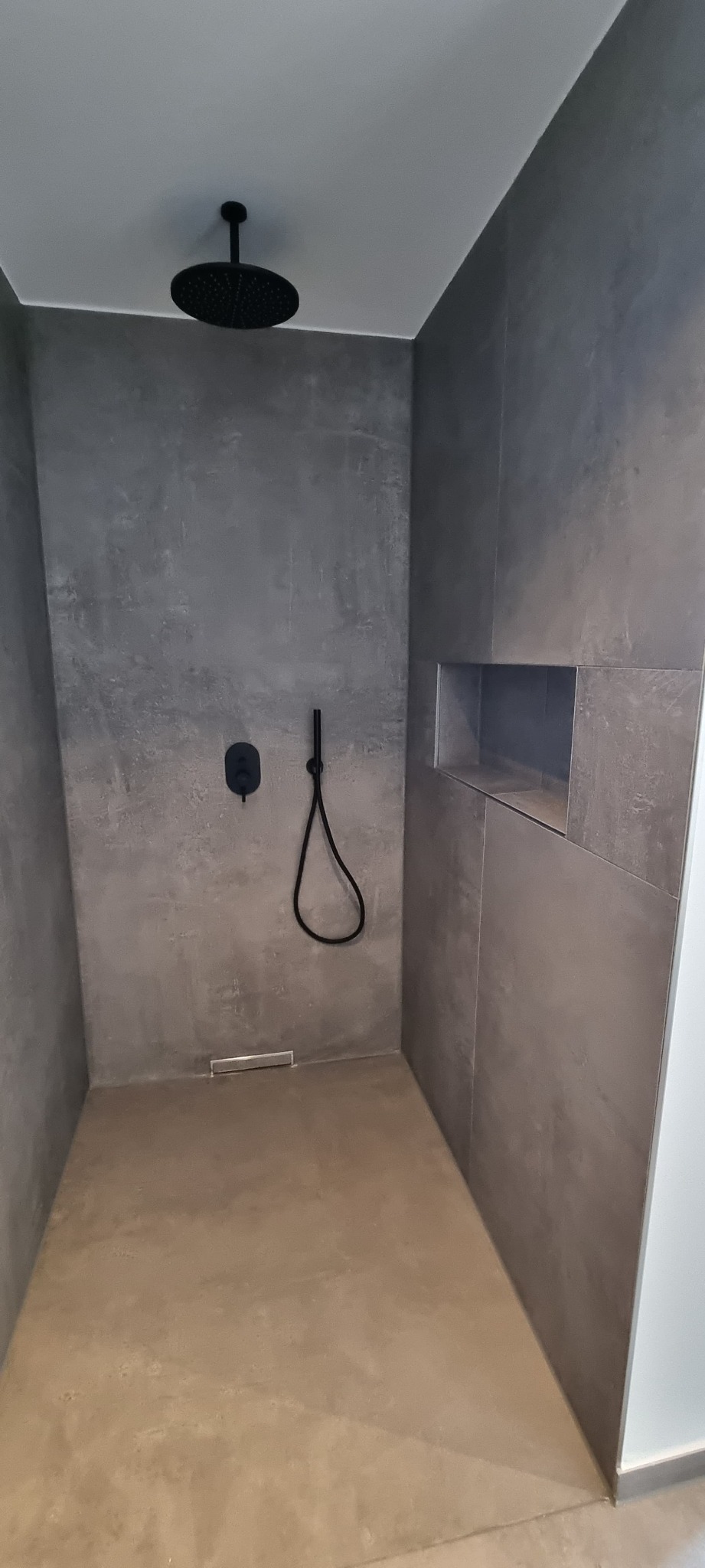 Nowoczesna kabina prysznicowa walk-in z szarymi płytkami imitującymi beton, czarną armaturą i wnęką na kosmetyki. Minimalistyczny design.