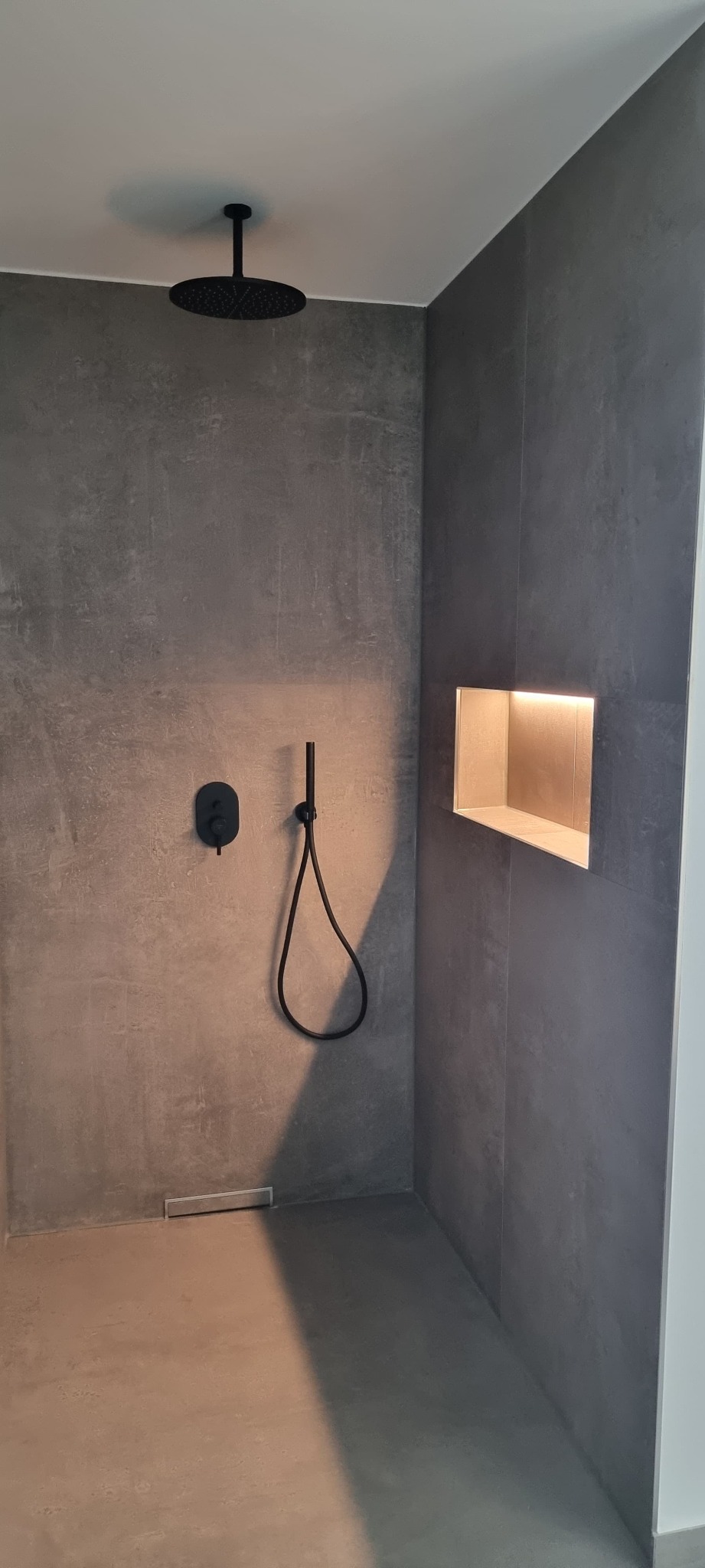 Minimalistyczna kabina prysznicowa z szarymi płytkami, czarną armaturą i wnęką z podświetleniem LED. Nowoczesny design, gładkie powierzchnie, deszczownica.