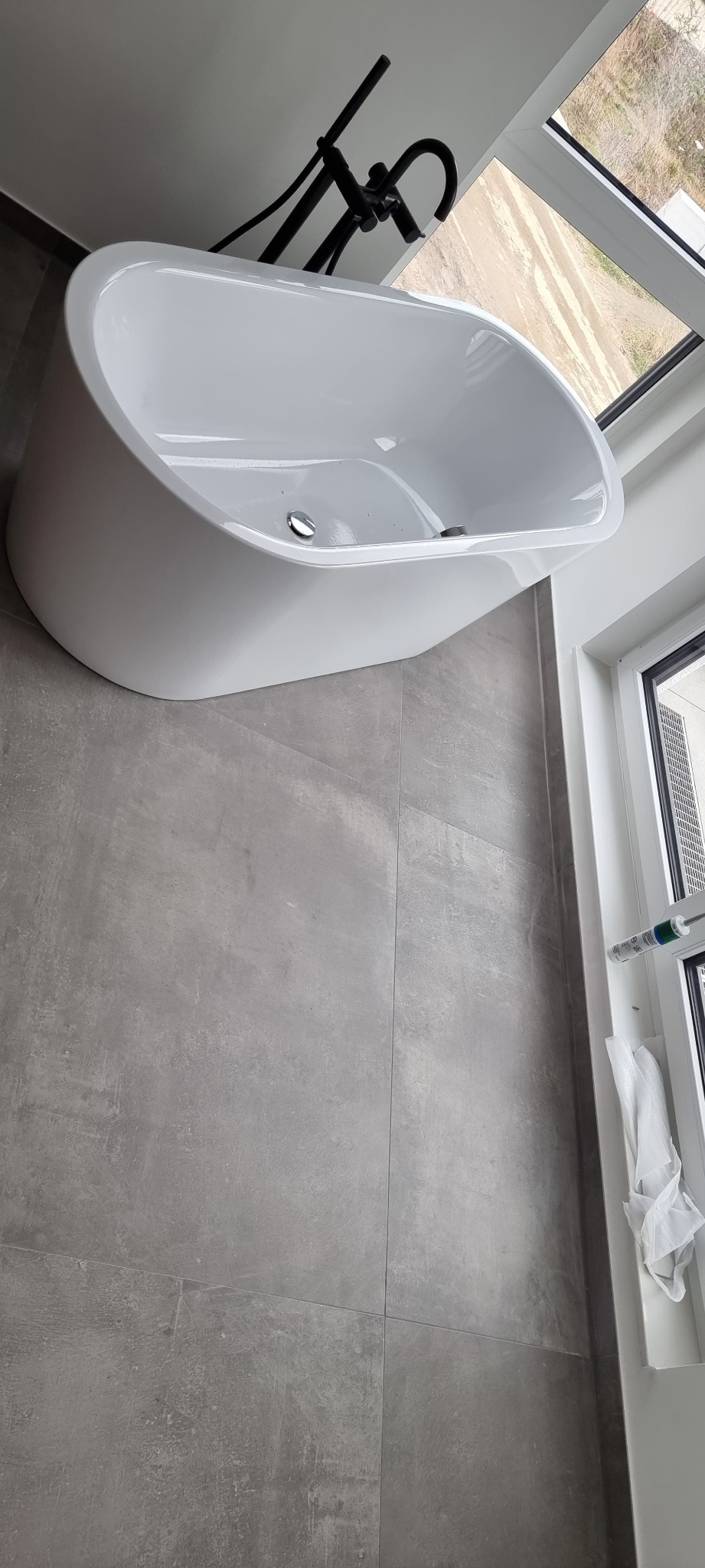 Nowoczesna, biała wanna wolnostojąca z czarną armaturą na tle szarych płytek i okna. Minimalistyczny design łazienki z elementami wykończeniowymi.