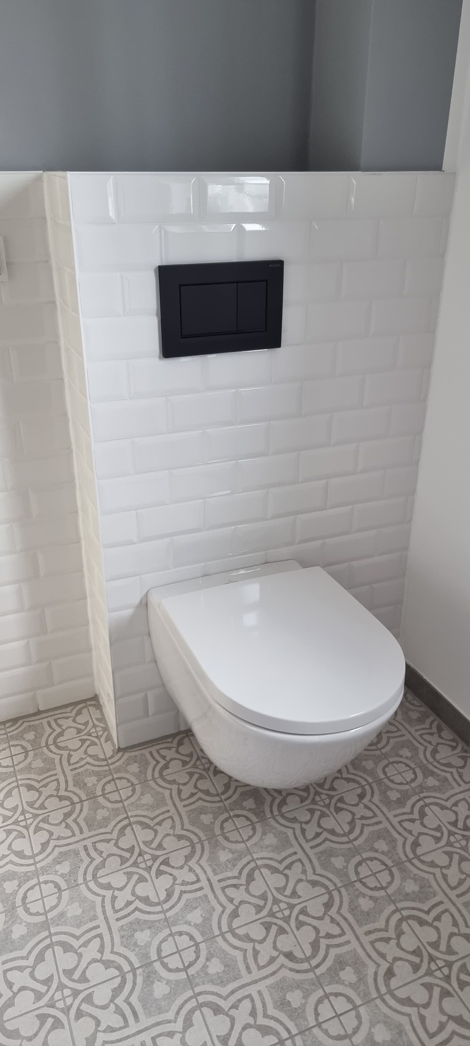 Wisząca toaleta z czarnym przyciskiem spłukującym na ścianie z białych, błyszczących płytek typu metro i wzorzystą szarą podłogą. Nowoczesna aranżacja łazienki.