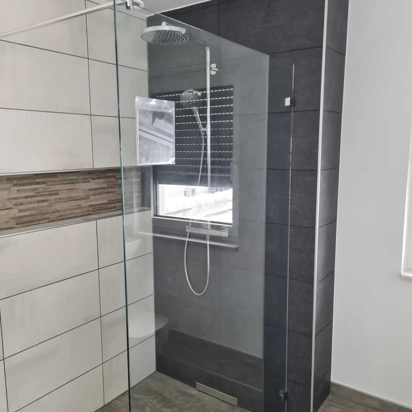 Nowoczesna kabina prysznicowa z deszczownicą, wykończona białymi i grafitowymi płytkami. Widoczne okno z roletą i dekoracyjny pas mozaiki. Minimalistyczny design.