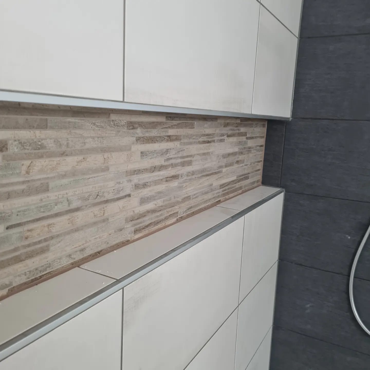 Wykończona wnęka z dekoracyjną, mozaikową okładziną w łazience. Białe, kwadratowe płytki ceramiczne z metalowymi listwami wykończeniowymi. Ciemnoszara ściana z natryskiem.