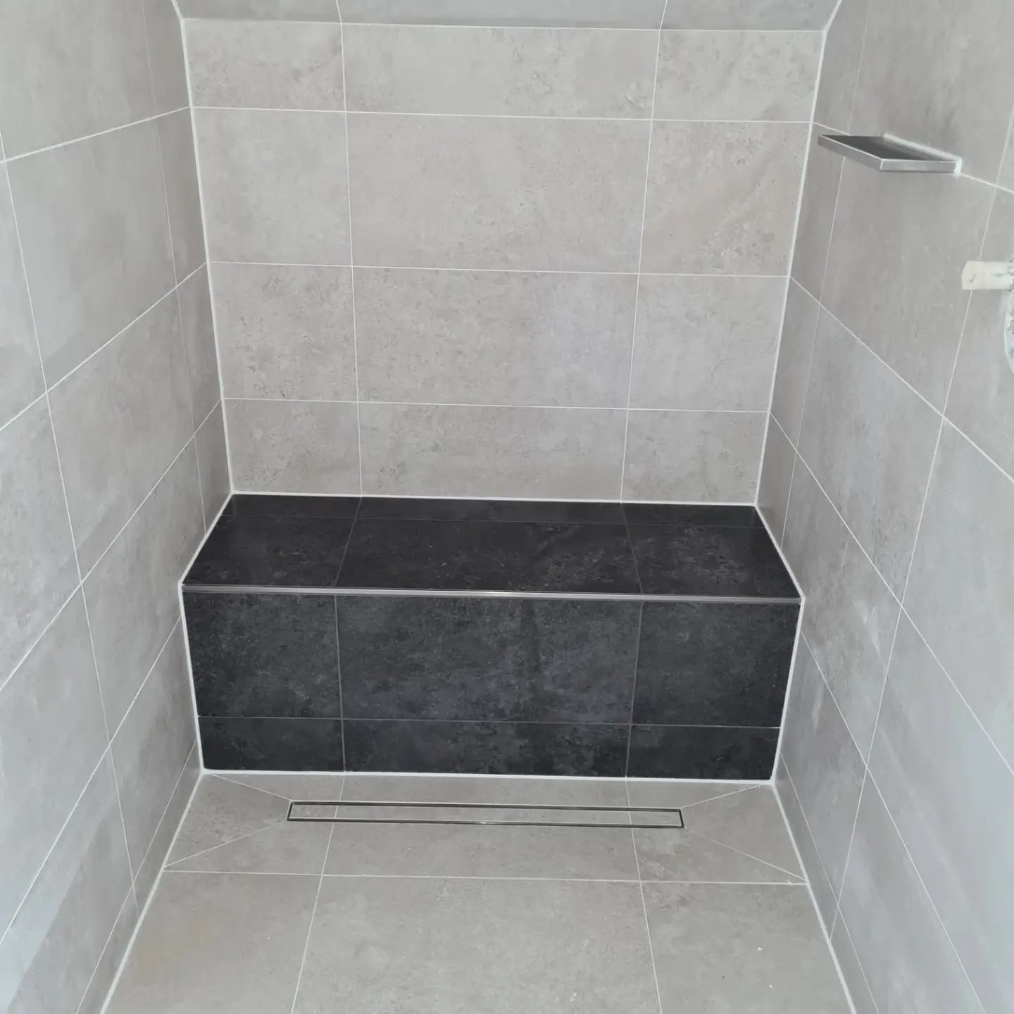 Wykończona kabina prysznicowa z siedziskiem obłożonym ciemnymi płytkami. Jasne, kwadratowe płytki na ścianach i podłodze. Widoczny odpływ liniowy.