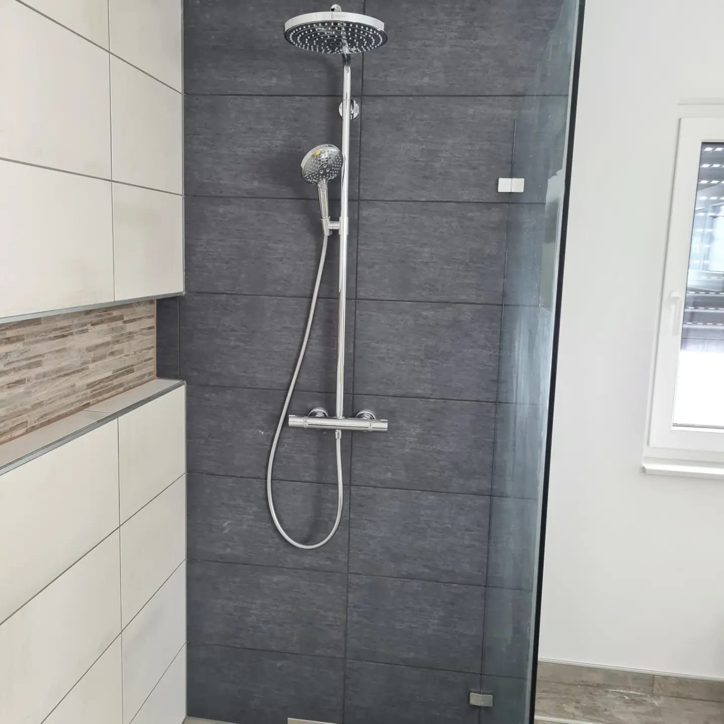 Nowoczesna kabina prysznicowa z deszczownicą i szarą płytką imitującą kamień, białe płytki i półka z mozaiką. Widoczna szyba kabiny i okno.