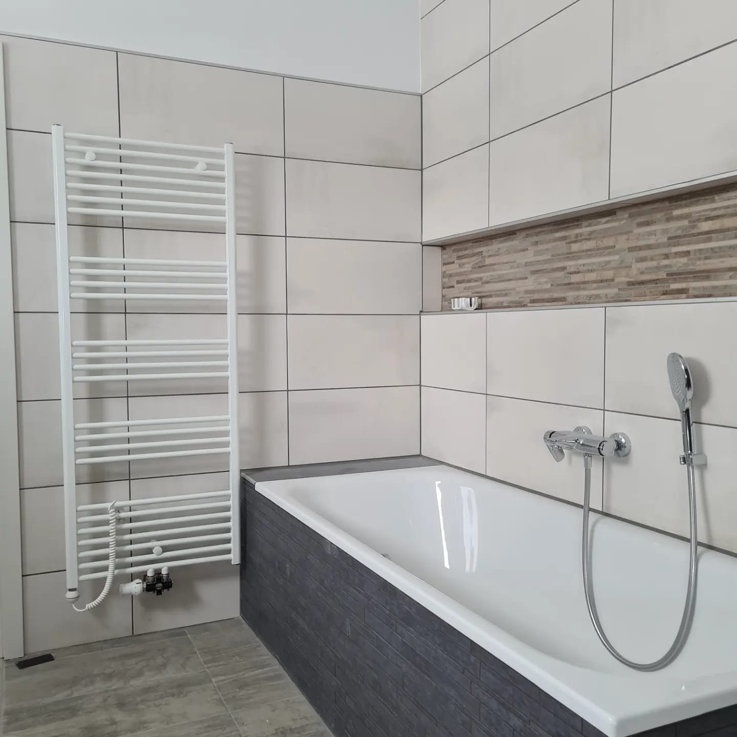 Wykończona łazienka z wanną obudowaną szarą mozaiką, grzejnikiem drabinkowym i jasnymi płytkami. W ścianie wnęka z dekoracyjną mozaiką. Nowoczesne baterie.